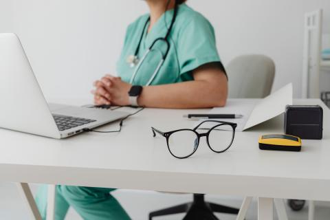 A person in scrubs sits behind their desk with a laptop and glasses in front of them